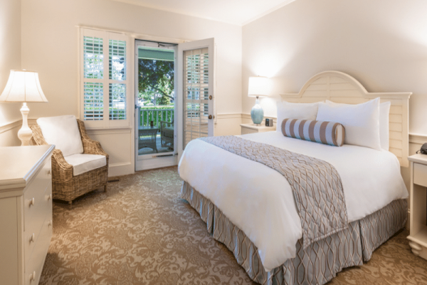 The image shows a neatly arranged bedroom with a bed, lamps, a chair, and a door leading to an outdoor view with greenery.