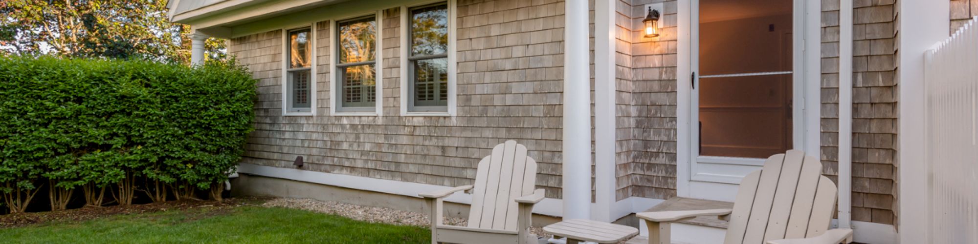 A cozy outdoor setting with two Adirondack chairs and a small table on a stone patio next to a shingled house, with a neatly trimmed lawn.