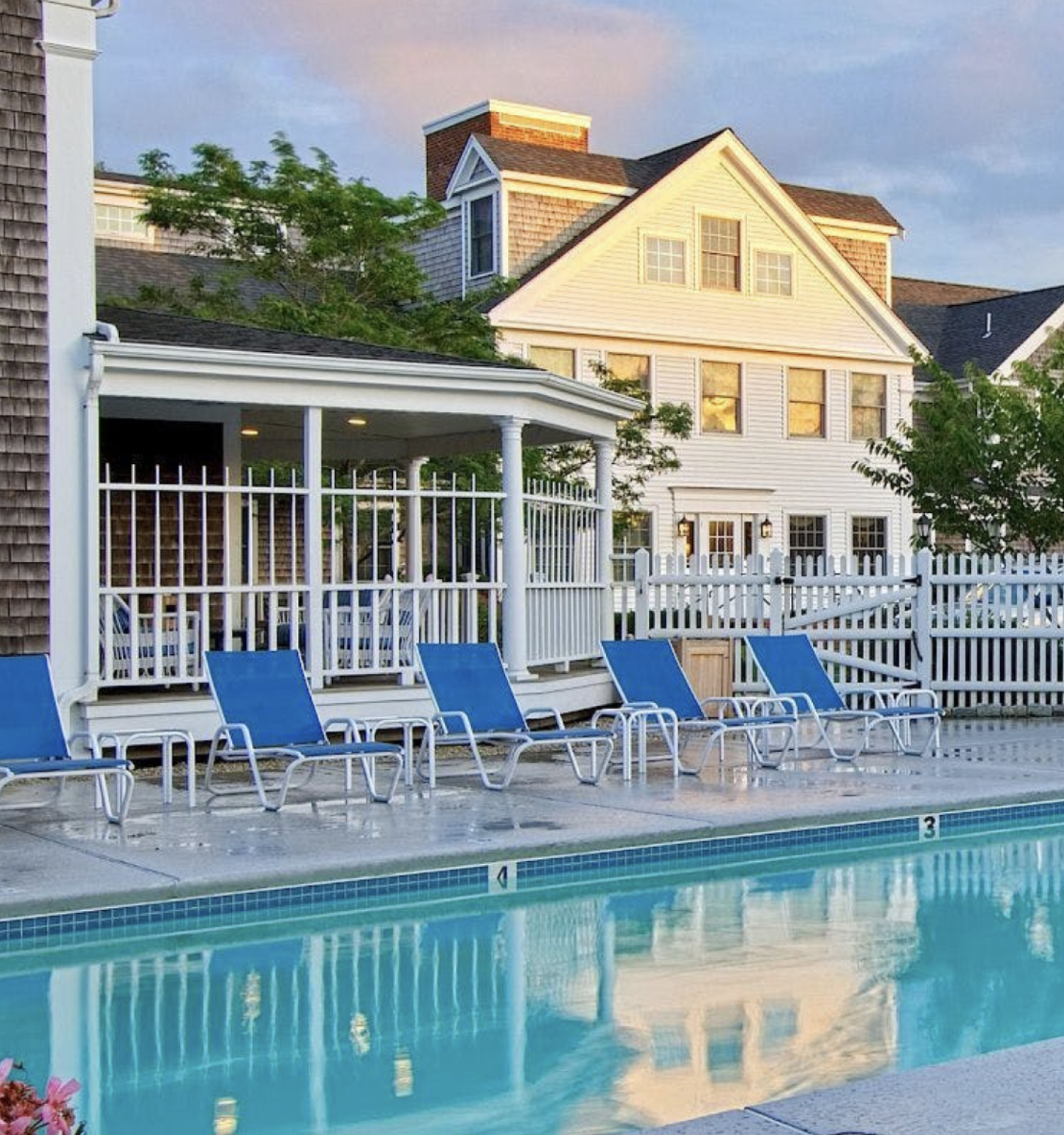 The image shows a serene pool area with blue lounge chairs and a white picket fence, adjacent to a classic residential building during sunset.