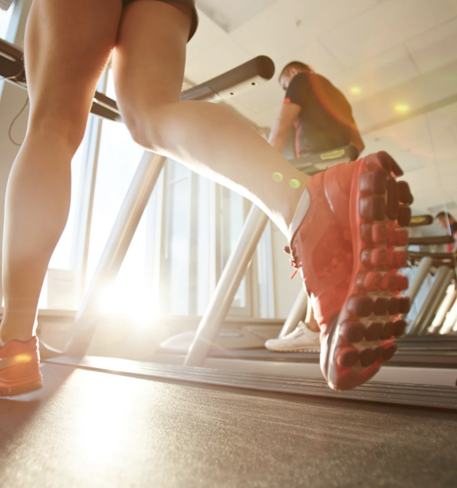 People running on treadmills in a gym with sunlight streaming through large windows. The focus is on the legs of a person wearing red running shoes.