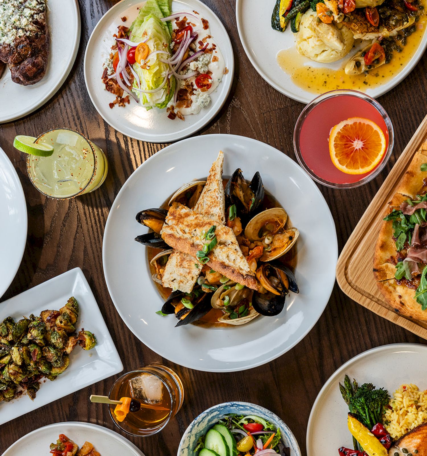 A table set with various dishes including pasta, seafood, salad, steak, and flatbread, along with colorful drinks and garnishes.