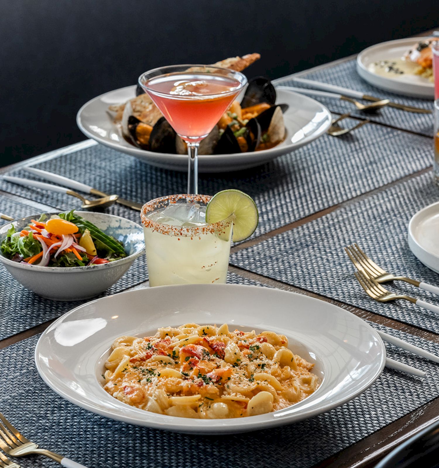 A table set with various dishes including pasta, salad, steak, shellfish, and colorful cocktails, all arranged on place settings and a woven table runner.