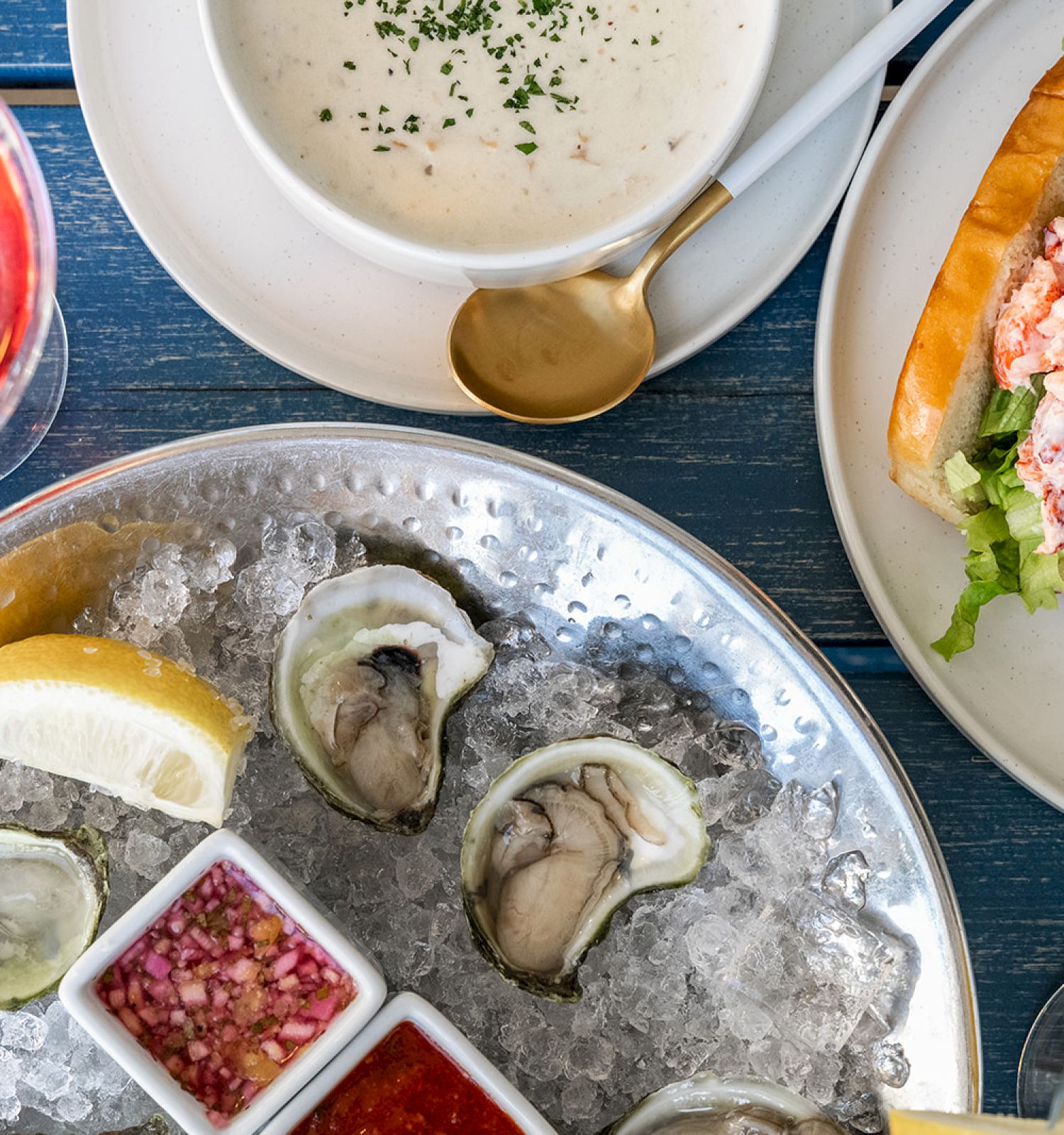 An overhead view of a seafood meal including oysters on ice, soup, a lobster roll, a pink drink, and condiments on a blue table.