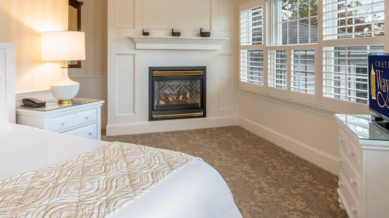 A cozy bedroom featuring a white bed, bedside table with a lamp, fireplace, dresser with a TV, and large shuttered windows.