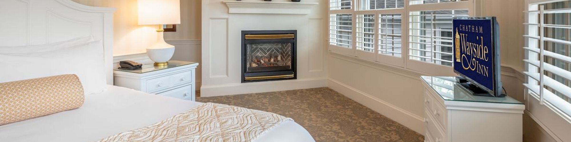 A cozy bedroom featuring a white bed, bedside table with a lamp, fireplace, dresser with a TV, and large shuttered windows.