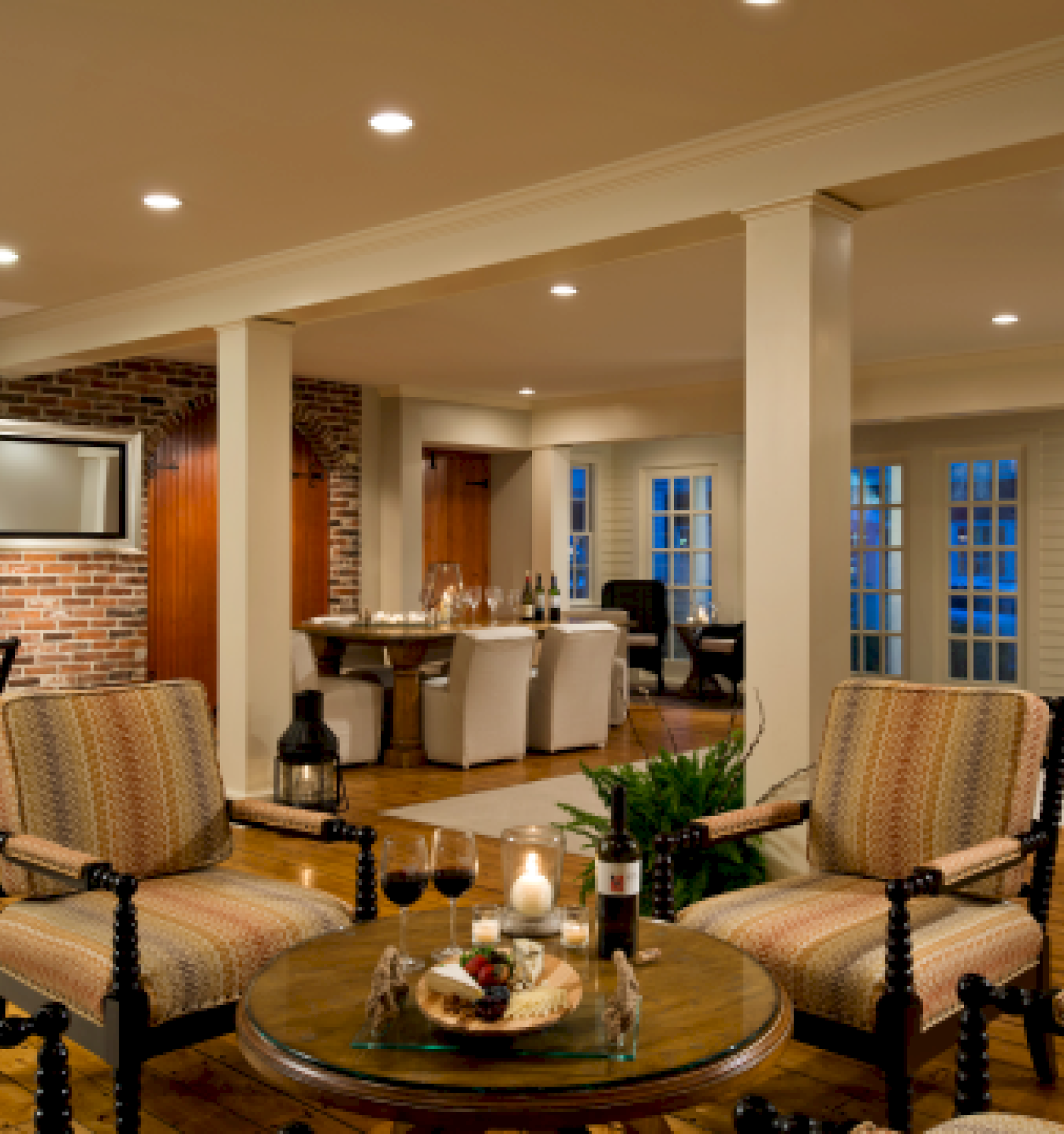 A cozy living room with armchairs, a round table with candles and wine, a grand piano, dining area, brick wall, and recessed lighting, always ending the sentence.