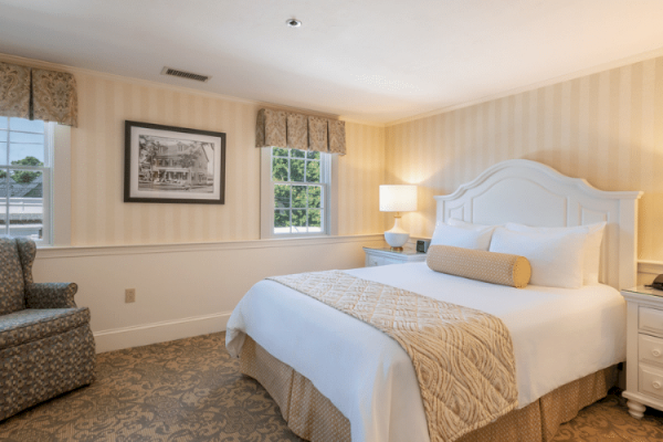 A cozy bedroom with a large bed, two lamps, a picture frame, a chair, and two windows, decorated in light, neutral tones.