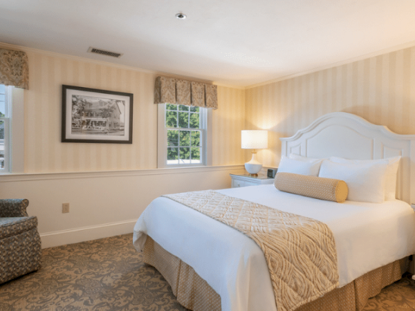 A cozy bedroom with a large bed, two lamps, a picture frame, a chair, and two windows, decorated in light, neutral tones.
