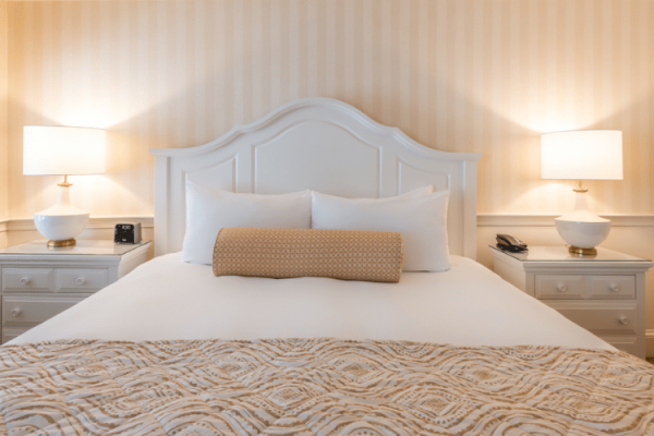 A neatly made bed with a white headboard, two side tables each with a lamp, a patterned blanket, and a bolster pillow in a beige-themed room.