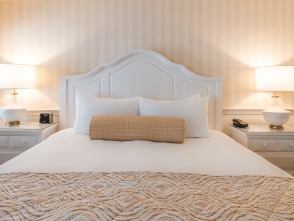 A neatly made bed with a white headboard, two side tables each with a lamp, a patterned blanket, and a bolster pillow in a beige-themed room.