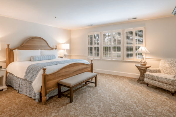 A cozy bedroom with a large bed, nightstands with lamps, a window with shutters, an armchair, a bench at the bed's foot, and a patterned carpet.