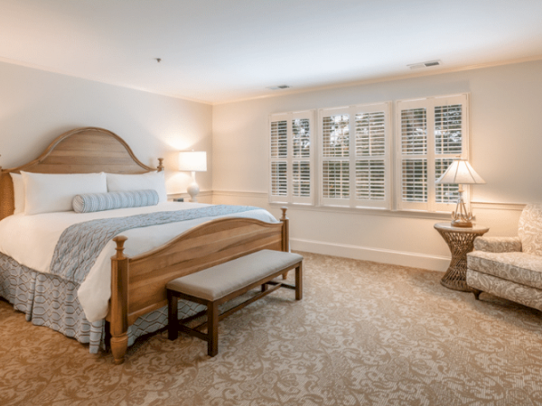 A cozy bedroom with a large bed, nightstands with lamps, a window with shutters, an armchair, a bench at the bed's foot, and a patterned carpet.