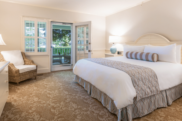 This image shows a cozy, well-lit bedroom with a large bed, two side tables, a wicker chair, and an open door leading to a balcony or patio area.