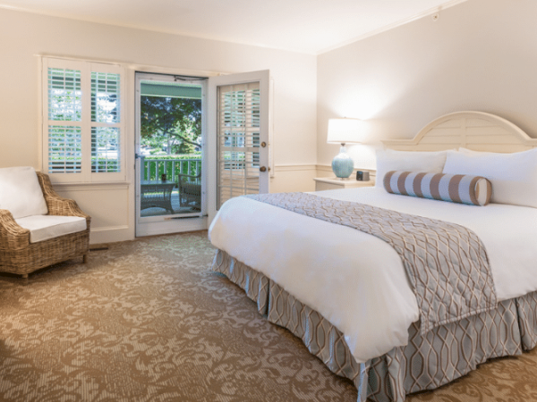 This image shows a cozy, well-lit bedroom with a large bed, two side tables, a wicker chair, and an open door leading to a balcony or patio area.
