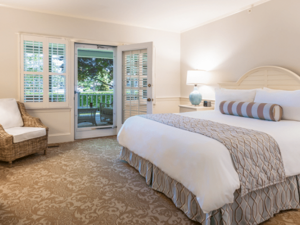This image shows a bright, cozy bedroom with a large bed, nightstands, armchair, and a door opening to a balcony or patio area.