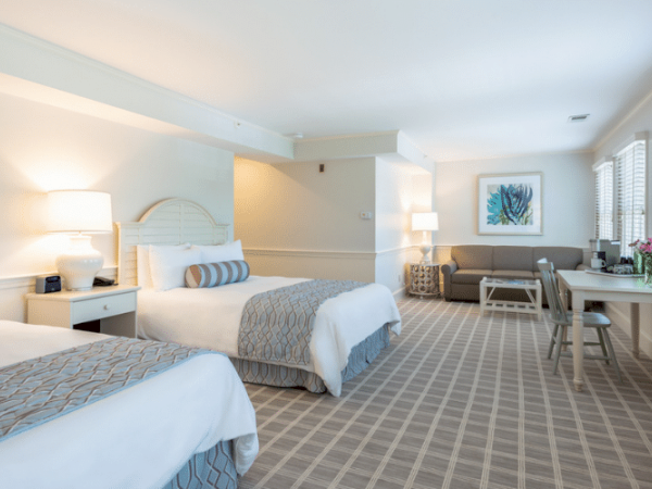 A hotel room with two beds, a desk, a TV, a sofa, and a large window providing natural light. The decor is light and modern.