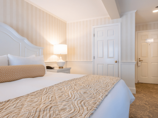 A bright hotel room features a large bed with pillows, a decorative bed runner, a bedside table with a lamp, and a door leading to the exit.