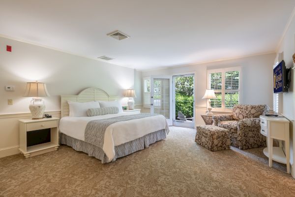 A spacious, well-lit bedroom with a large bed, two bedside tables, a cozy sitting area with chairs, a TV on the wall, and a door leading outside.