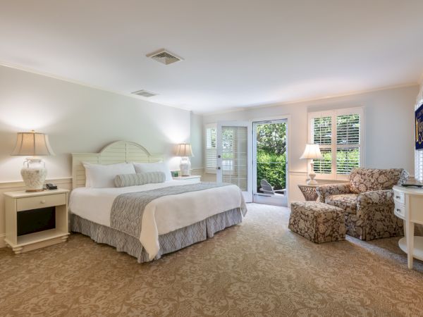 A spacious, well-lit bedroom with a large bed, two bedside tables, a cozy sitting area with chairs, a TV on the wall, and a door leading outside.