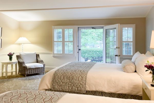 A cozy well-lit bedroom features two neatly made beds, nightstands with flowers, a white armchair, framed art, and a door leading outside.