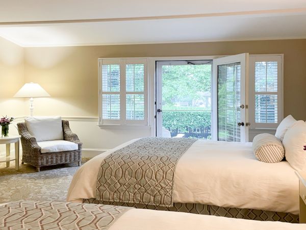 A cozy well-lit bedroom features two neatly made beds, nightstands with flowers, a white armchair, framed art, and a door leading outside.