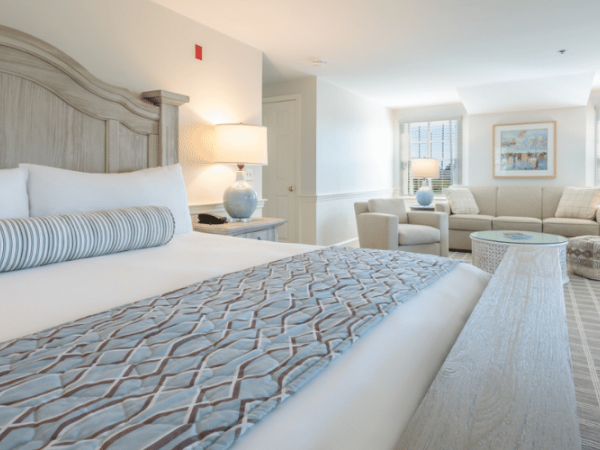 A bright, cozy hotel room with a king-sized bed, elegant blue and white decor, a sitting area with a sofa, armchairs, and two lamps by the window.