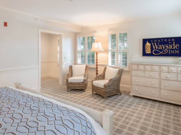 A cozy bedroom with a bed, dresser, wicker chairs, and a sign for "Chatham Wayside Inn" on the wall. The room is decorated in neutral tones.