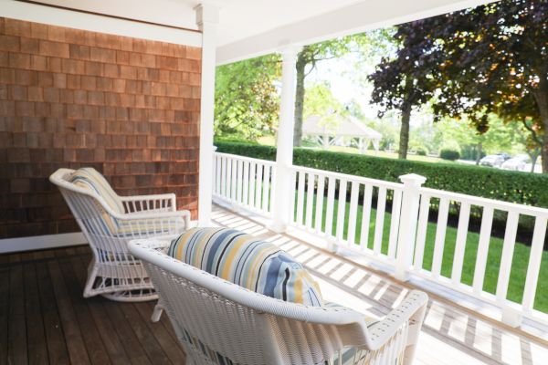 A cozy porch with two wicker chairs with striped cushions, overlooking a well-maintained garden and lawn through white railings, during a sunny day.