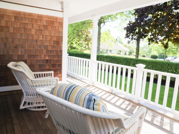 A cozy porch with two wicker chairs with striped cushions, overlooking a well-maintained garden and lawn through white railings, during a sunny day.