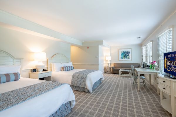 The image shows a neatly arranged hotel room featuring two beds with blue-gray accents, a couch, wall art, a TV, and a desk.