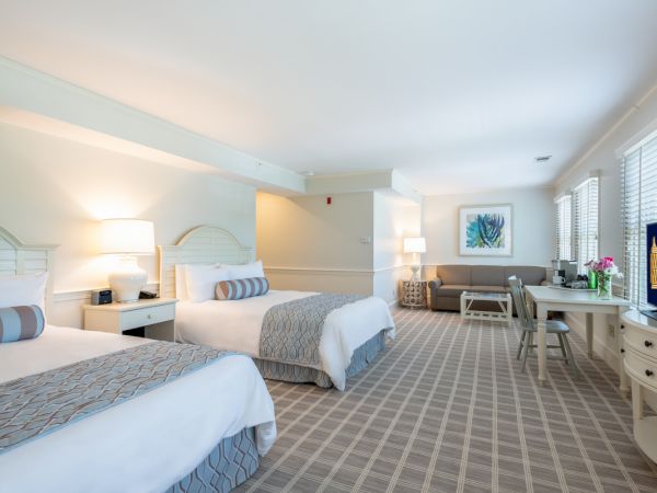 The image shows a neatly arranged hotel room featuring two beds with blue-gray accents, a couch, wall art, a TV, and a desk.