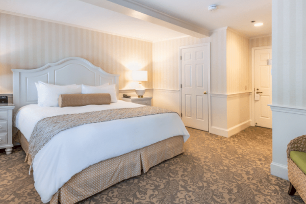 The image shows a well-lit hotel room with a bed, nightstands, lamps, and a door leading to the hallway, carpeted with a patterned floor.