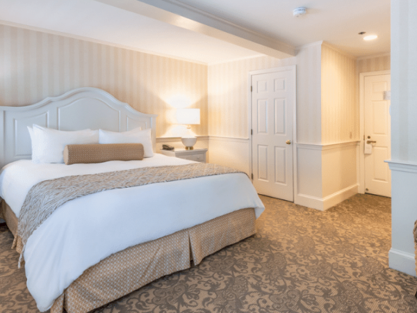 The image shows a well-lit hotel room with a bed, nightstands, lamps, and a door leading to the hallway, carpeted with a patterned floor.