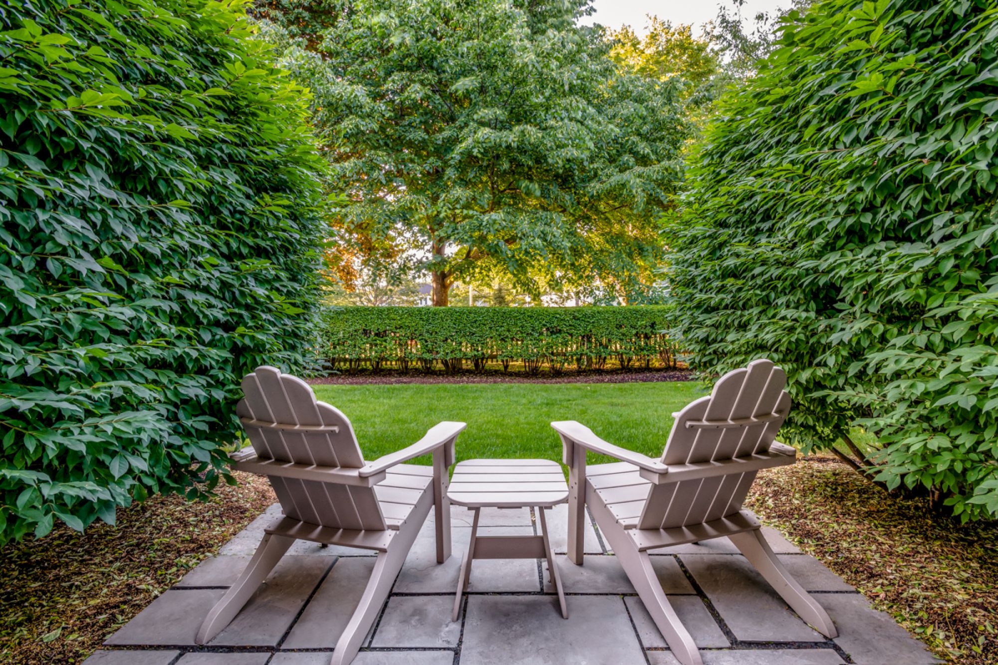 The image shows two Adirondack chairs with a small table in between, placed on a patio surrounded by lush greenery and trees in the background.