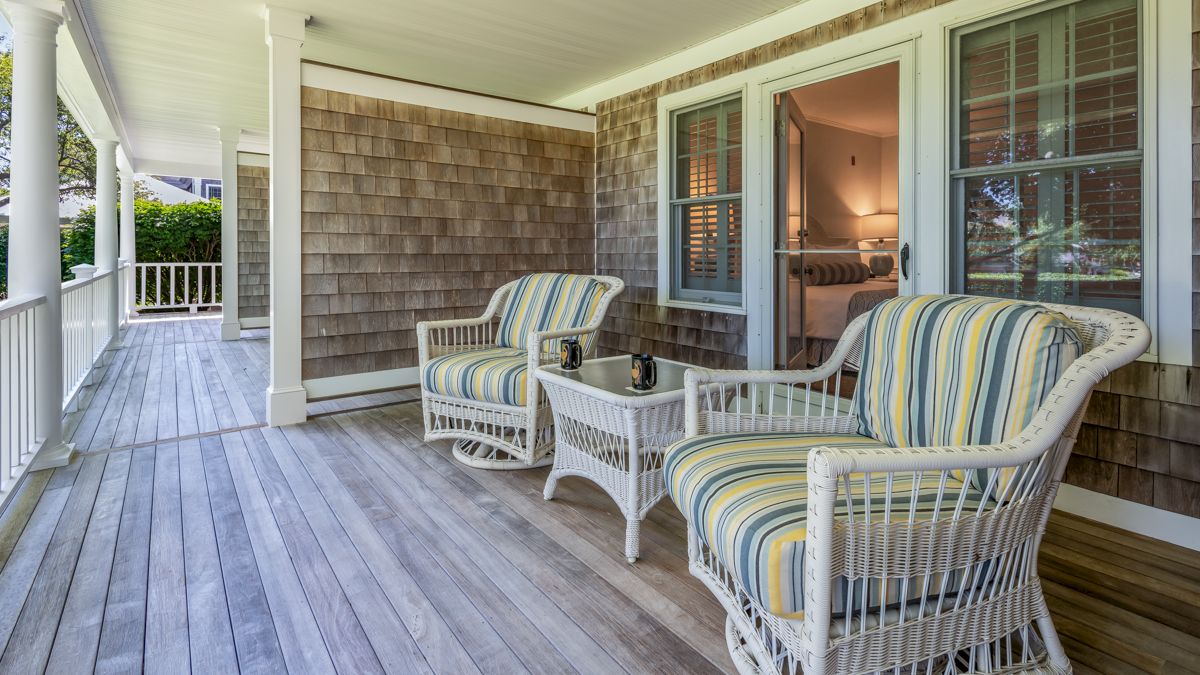 A cozy porch with wicker chairs, striped cushions, a small table, and two mugs, next to a house with shingle siding and a glass door.