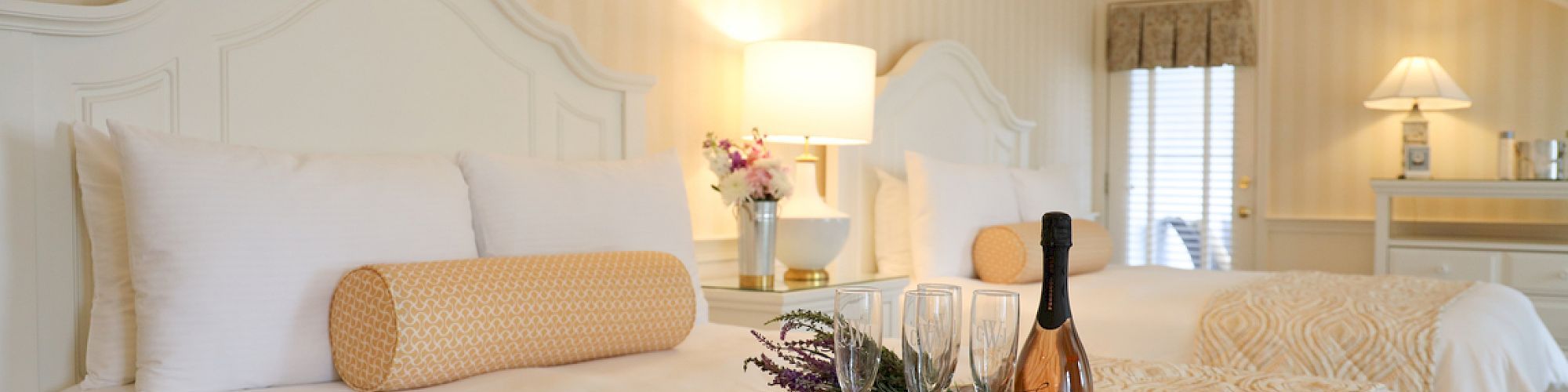 A cozy hotel room with two white beds, a tray with a bottle, glasses, plant, and a room lamp on a bedside table.