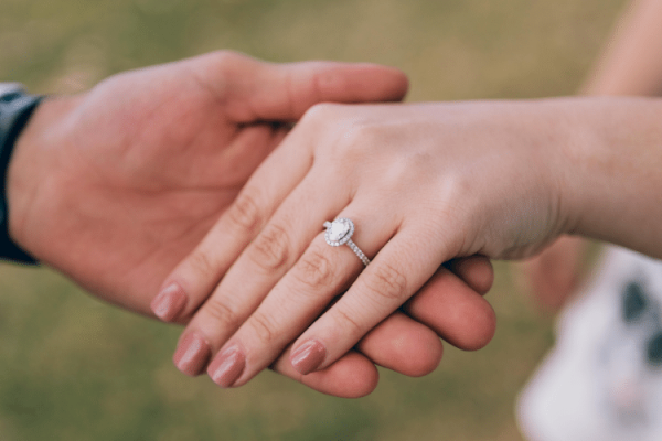 The image shows two hands holding each other, with one hand wearing a diamond engagement ring.