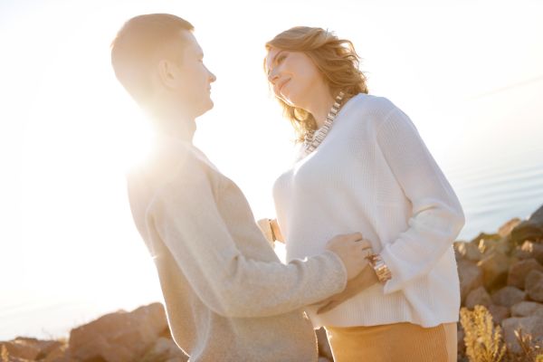 A couple stands near water with sunlight in the background, the man touches the woman's belly as they both smile.