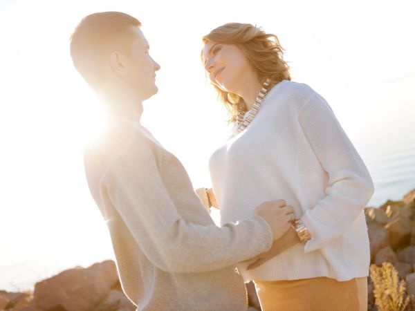 A couple stands near water with sunlight in the background, the man touches the woman's belly as they both smile.