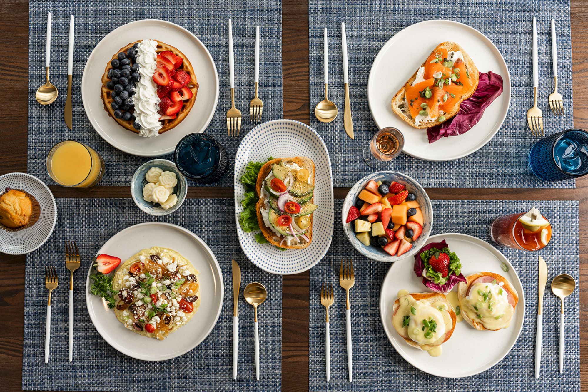 An overhead view of a well-organized breakfast spread with a variety of dishes, including fruit, waffles, toast, eggs benedict, and various drinks.