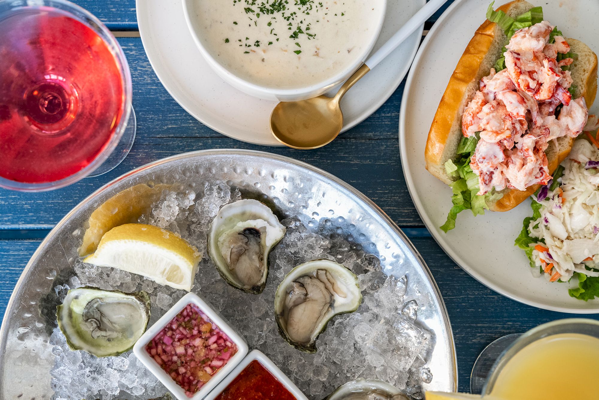 The image shows a meal with oysters on ice, a lobster roll, chowder, a glass of red wine, and various condiments on a blue table.