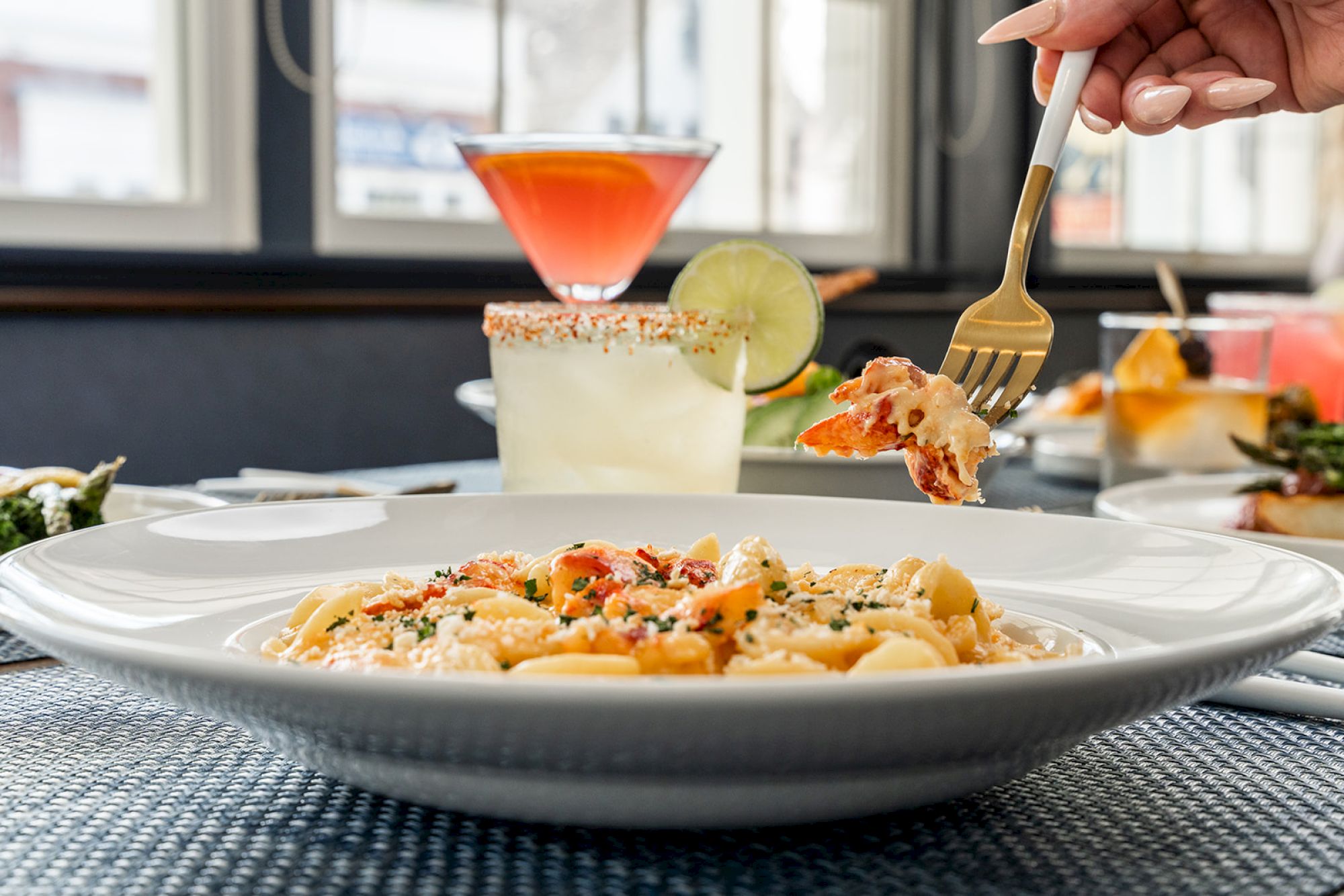 Pasta dish with shrimp on a fork, surrounded by cocktails and a well-lit dining area in the background.