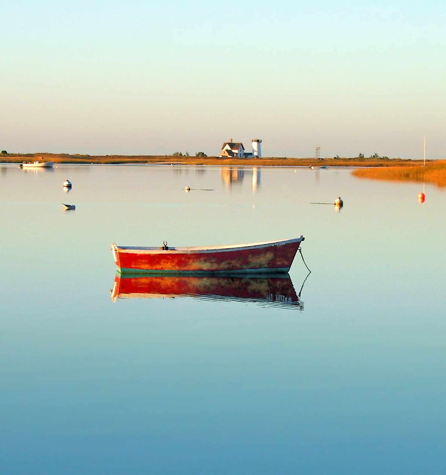 A lone red boat floats on calm water with buoys and a distant shoreline featuring a small house and trees under a clear sky.