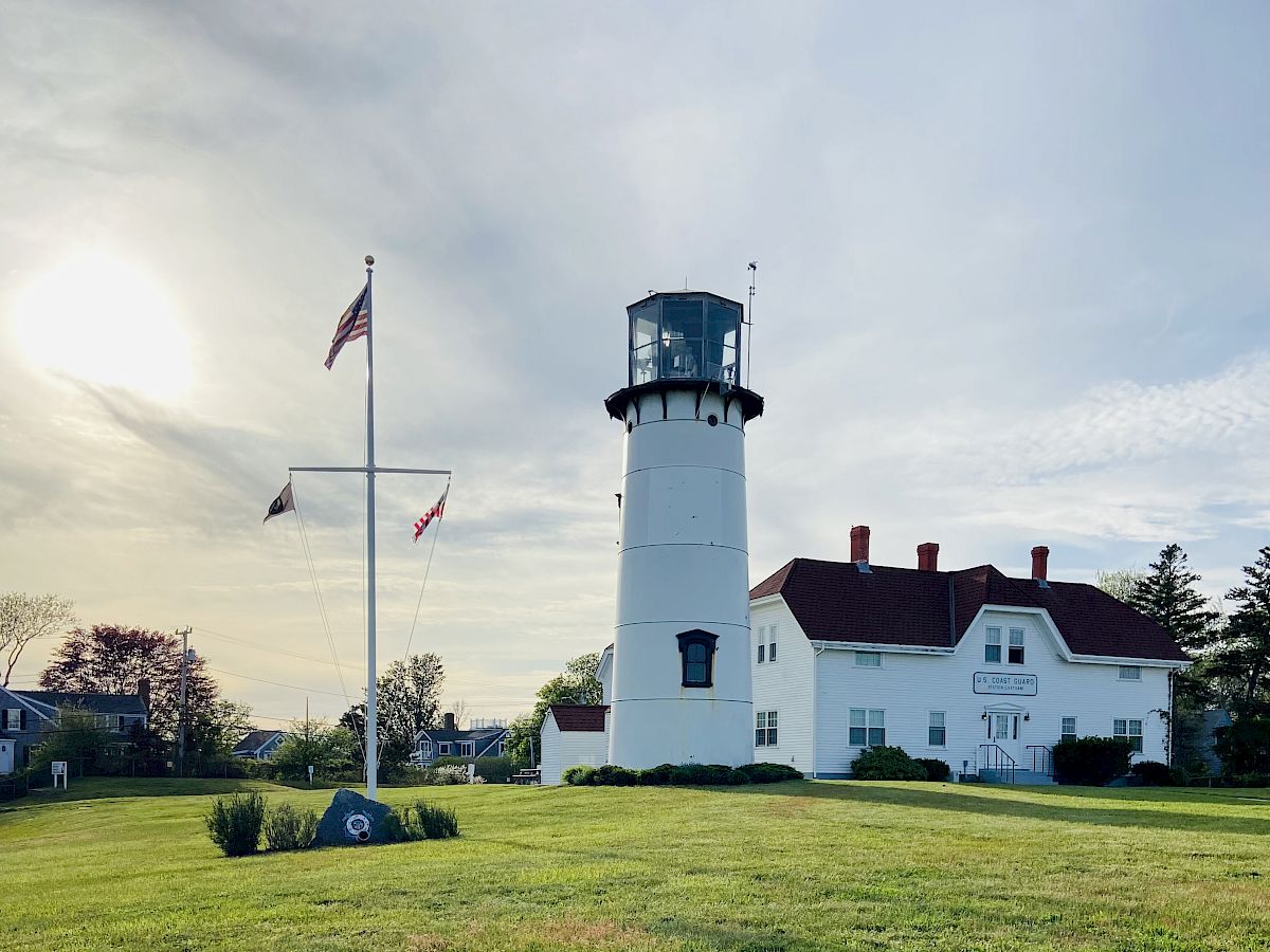 A picturesque lighthouse and its adjoining building are surrounded by a well-maintained grassy field, with a flagpole nearby under a serene sky.