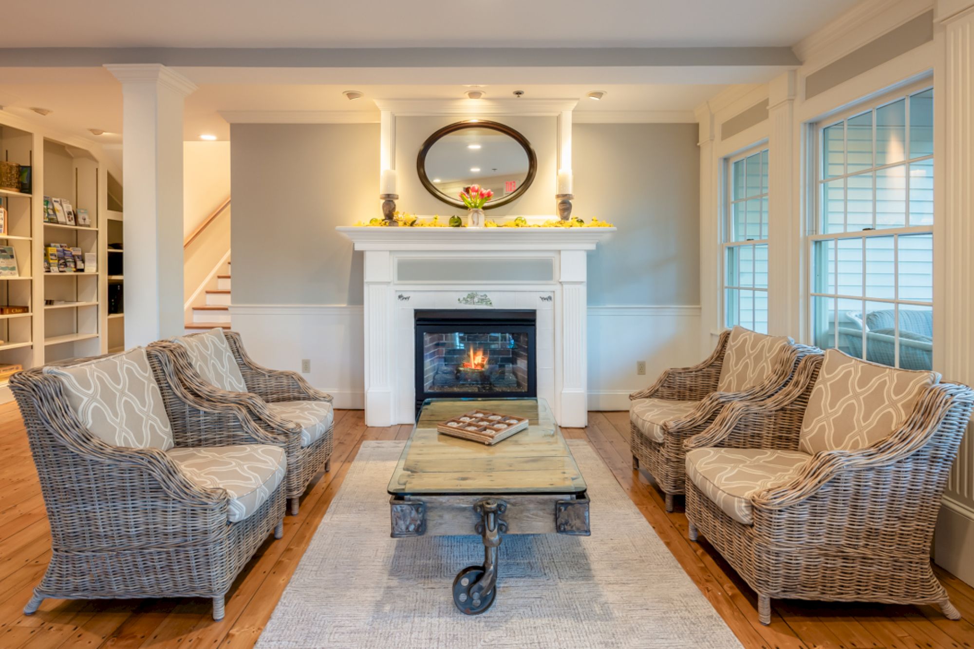 A cozy living room with four woven armchairs around a wooden table. There's a fireplace with a mirror above it and shelves in the background.