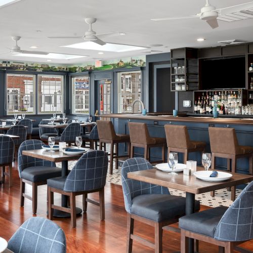 The image shows a modern, well-lit restaurant bar with a sleek design, featuring navy blue chairs, polished wooden floors, and a stocked bar in the background.