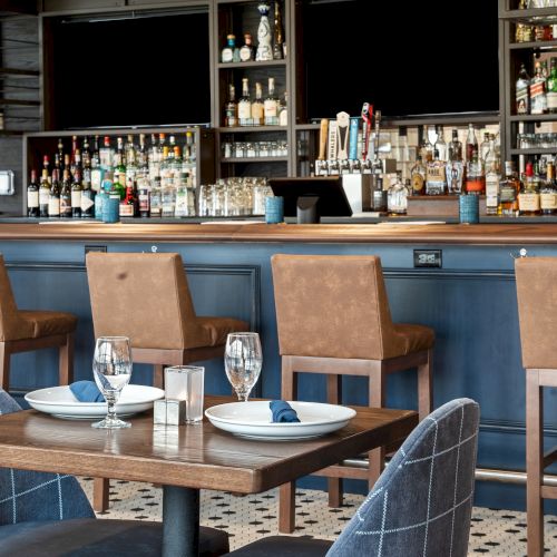 The image shows a cozy bar with a modern design, featuring bar stools, a variety of bottles, glasses, and two tables set for dining.