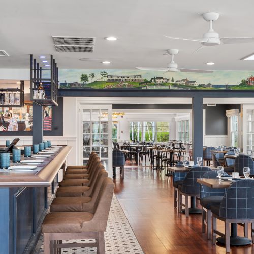 A modern, spacious restaurant interior with a bar area on the left, cushioned chairs, and tables arranged neatly, featuring natural light from large windows.