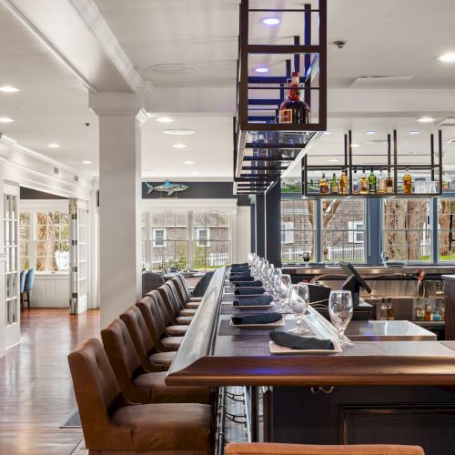 This image features a modern, well-lit bar area with leather stools lined up along a wooden counter. Shelves display various spirits.