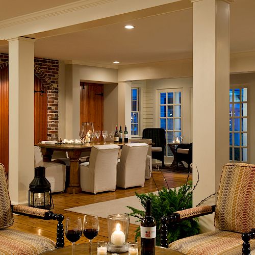 A cozy, elegant living room features patterned armchairs, wine glasses, candles, exposed brick walls, white columns, a dining area, and a black grand piano.